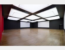 Großer, moderner Ausstellungsraum mit dunklen Wänden, hölzernem Boden und großen rechteckigen LED-Deckenleuchten. Zwei leere weiße Tafeln hängen an den Wänden, eine rote Wand befindet sich links.