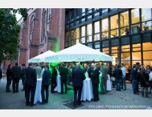 Außenbereich einer Veranstaltung mit mehreren weißen Pavillons und Stehtischen, umgeben von Menschen in formeller Kleidung. Im Hintergrund eine Fassade mit roten Backsteinen und hohen Fenstern sowie ein modernes Gebäude mit Glas- und Metallfront und beleuchteten Innenräumen.