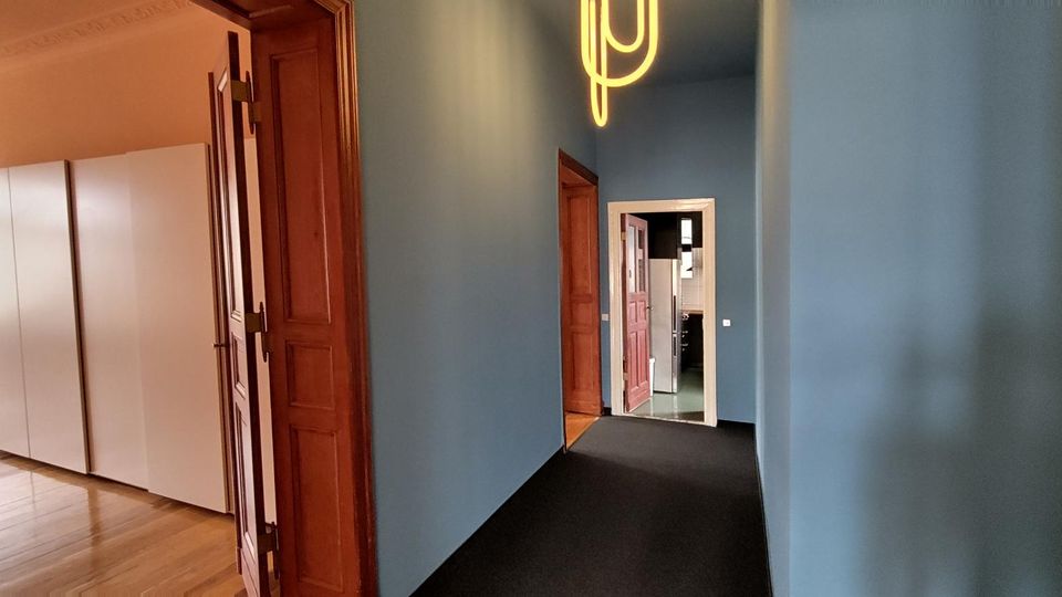 Schmaler Flur mit blau gestrichenen Wänden, dunklem Teppichboden und hohen, braunen Holztüren. Am Ende führt ein geöffneter Durchgang in einen weiteren Raum, an der Decke hängt eine moderne Leuchte.