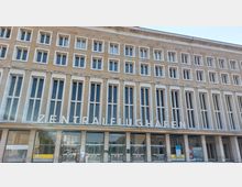 Fassade eines historischen Gebäudes mit der Aufschrift "Zentralflughafen" in großen, weißen Buchstaben. Die Architektur zeigt rechteckige Fenster in symmetrischer Anordnung, mit einem Eingangsbereich aus Glas im Erdgeschoss.