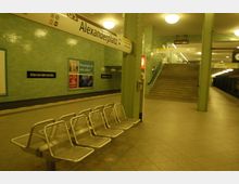 U-Bahn-Station mit der Beschilderung „Alexanderplatz“, grün gefliesten Wänden, Metallbänken im Wartebereich sowie einer Treppe, die zur höheren Ebene führt. Die Umgebung ist hell ausgeleuchtet, und ein Bahnsteig ist im Hintergrund sichtbar.