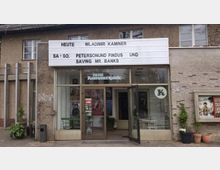 Außenansicht eines kleinen Kinos namens "Neue Kammerspiele" mit einem weißen Reklameschild, das aktuelle Veranstaltungen und Filme ankündigt. Der Eingangsbereich hat große Glasfenster, durch die Stühle und Tische sichtbar sind, und links vom Eingang stehen Pflanzkübel.