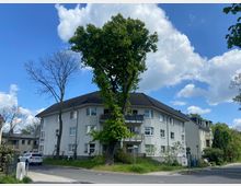 Mehrfamilienhaus an einer Straßenecke mit hellgrauer Fassade und dunkelgrauem Satteldach. Vor dem Gebäude steht ein großer Baum, umgeben von Grünfläche, während der Himmel klar und teils bewölkt ist.