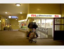 Eingangsbereich einer U-Bahn-Station mit gelben Wandfliesen. Rechts ein beleuchteter Waschsalon mit der Aufschrift „fit Waschcenter“, davor ein Ständer mit Taschen; links führen Rolltreppen in den Untergrund, eine große Uhr hängt von der Decke.