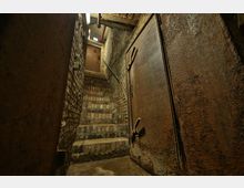 Blick in einen alten, gemauerten Keller mit schmaler Treppe aus Ziegelsteinen. Rechts im Bild befindet sich eine schwere, rostige Metalltür, die Raum und Atmosphäre industriell wirken lässt.