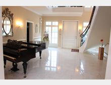 Elegantes, helles Foyer mit cremefarbenen Wänden, glänzendem Marmorboden, einer geschwungenen Treppe mit schwarzem Geländer, einem großen schwarzen Flügel und einem dekorativen Spiegel an der Wand. Im Hintergrund eine weiße Tür und bodentiefe Fenster, daneben eine große Vase mit Blumen.