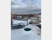 Schneebedeckte Dachterrasse mit einfachen Gartenmöbeln, darunter eine Liege, ein Stuhl und ein Tisch. Im Hintergrund ist eine Stadtlandschaft mit verschneiten Dächern und einem bewölkten Himmel zu sehen.
