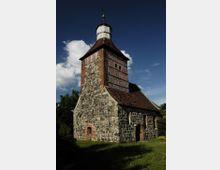 Historische Dorfkirche aus Feld- und Backsteinen mit einem steilen Ziegeldach, Fachwerkelementen im Turm und einem achteckigen Dachaufbau mit Wetterfahne, umgeben von grüner Wiese und Bäumen unter blauem Himmel.