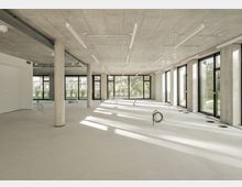 Innenraum eines modernen Rohbaus mit Betonwänden, -decken und -säulen. Große Fensterfronten lassen viel Tageslicht herein und geben den Blick auf eine grüne Außenanlage frei.