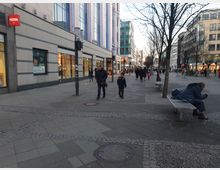Belebte Fußgängerzone mit Geschäften auf beiden Seiten, darunter eine Filiale von HEMA. Gehweg mit gepflasterten Mustern, Bäumen und Bänken, auf denen Menschen sitzen; im Hintergrund weitere Passanten und moderne Gebäude.