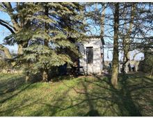 Kleines weißes Gebäude in einem Garten, umgeben von Nadelbäumen und Rasen. Im Vordergrund steht ein Tisch mit Stühlen, teilweise im Schatten der Bäume.