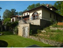 Ein zweistöckiges Gebäude im mediterranen Stil mit hellen Putzwänden, großen Rundbogenfenstern und einem steinernen Untergeschoss. Das Haus ist von Gartenanlagen und Rasenflächen umgeben, mit einer Terrasse, die von einem Metallgeländer begrenzt wird, und liegt in einer bewaldeten Umgebung.