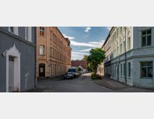 Straßenszene mit historischen Wohngebäuden, darunter ein graues Haus links und ein hellgelbes Gebäude rechts, die eine enge gepflasterte Straße säumen. In der Mitte steht ein Baum, und geparkte Autos reihen sich entlang der Straße bis zu einem Bereich mit kleineren Häusern im Hintergrund.