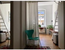 Ein heller Raum mit Holzboden und einer großen Fensterfront, durch die Tageslicht einfällt. Im Vordergrund steht ein grüner Stuhl vor einem weißen Vorhang, seitlich sind Leitern angelehnt, und im Hintergrund befindet sich ein Schreibtisch mit Pflanzen.