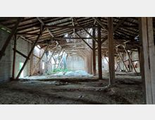 Innenraum eines verfallenen Gebäudes mit freiliegendem Holzdachstuhl, Backsteinwänden und gestützten Holzbalken. Der Boden ist uneben und mit Schutt bedeckt, während Licht durch beschädigte Stellen im Dach einfällt.