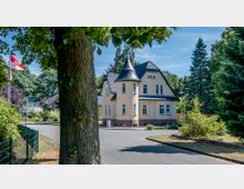 Zweistöckiges cremefarbenes Haus mit einem runden Erkerturm und dunklem Schieferdach, umgeben von Bäumen und Grünflächen. Im Vordergrund ein Baumstamm sowie Straßenschilder und Flaggen am Straßenrand.