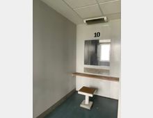 Kleiner, karger Innenraum mit grauen Wänden und abgehängter Decke, an der Stirnwand ein Fenster mit der Nummer „10“ darüber und einer langen, schmalen Ablage darunter. Im Raum steht ein fester, quadratischer Hocker auf einem Metallfuß; der Boden ist dunkel und glatt.