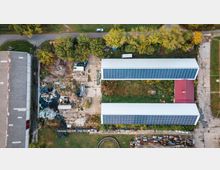 Luftaufnahme eines Industrie- oder Landwirtschaftsgeländes mit zwei langen Gebäuden, deren Dächer vollständig mit Solarmodulen bedeckt sind. Dazwischen befinden sich eine Grünfläche und ein Bereich mit Lagerplätzen und verstreutem Schutt, umgeben von Bäumen und einer Straße.