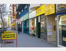 Straße mit einer Reihe kleiner Geschäfte und Praxen, darunter ein Fahrschulbüro mit gelben Schildern, ein Fotogeschäft und weitere Läden mit bunten Fassaden. Links stehen Bäume, und einige Passanten sind auf dem Gehweg unterwegs.