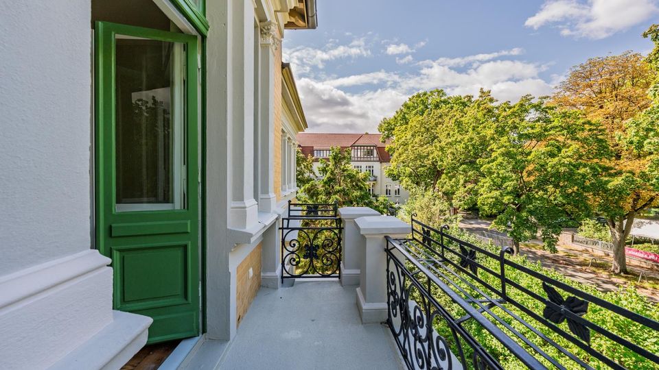 Balkon an einer historischen Fassade mit weiß verzierten Stuckelementen, grüner Balkontür und schwarzem schmiedeeisernen Geländer. Blick über hohe Laubbäume auf ein gegenüberliegendes Gebäude mit rotem Dach unter blauem Himmel.