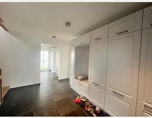 Flur eines Wohnhauses mit weißen Einbauschränken an der rechten Wand, dunklem Bodenbelag aus Fliesen und einem Durchgang, der zu einem Bereich mit Tageslicht führt. Auf dem Boden stehen Schuhe vor den Schränken, und links ist ein Teil einer Holztreppe sichtbar.