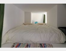 Schlafnische in einem minimalistischen Raum mit niedriger Decke und weißen Wänden, darin ein großes Bett mit weißer Bettwäsche. Am Fußende liegt ein gemusterter Teppich; hinten sind ein grüner Vorhang und ein blauer Globus zu sehen.