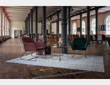 Großer, heller Saal mit hohen Decken und gusseisernen Säulen, eingerichtet mit Stühlen, Sesseln und kleinen Tischen auf einem Teppich. Im Hintergrund sind Holzstühle in Reihen angeordnet, große Fenster mit Rundbögen lassen Tageslicht herein.