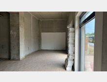 Ein leerer Innenraum mit Betonwänden und Rohdecke, hellem Laminatboden und einem großen Fenster auf der rechten Seite. Im Vordergrund sind gestapelte Styroporplatten zu sehen, und außerhalb des Fensters ist ein Gehweg sowie ein gelber Linienbus erkennbar.