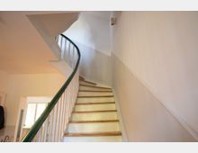 Innenansicht eines Treppenhauses mit einer schmalen, nach oben führenden Holztreppe und einem geschwungenen Geländer mit dunklem Handlauf und weißen Stäben. Weiße Wände und eine Deckenleuchte prägen den hellen Flur, unten ist ein Türdurchgang zu sehen.