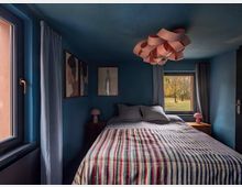 Ein kleines Schlafzimmer mit blauen Wänden und zwei Fenstern, die natürliches Licht hereinlassen. Das Zimmer zeigt ein Bett mit gestreiftem Bettüberwurf, zwei Nachttischen mit Pilzlampen und modernen Wandbildern, sowie eine auffällige Deckenleuchte in geschwungener Form.