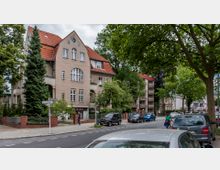 Wohnstraße mit einer mehrstöckigen Altbauvilla im Vordergrund, die ein rotes Ziegeldach und helle Putzfassade aufweist. Rechts daneben sind weitere Wohnhäuser mit Balkonen zu sehen, und die Straße ist von geparkten Autos sowie Bäumen gesäumt.