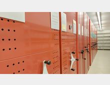 Rote Rollregale in einem Archivraum mit Kurbeln zur Bedienung. Rechts sind leere weiße Regalböden zu sehen, die Beleuchtung erfolgt durch lange Deckenleuchten.