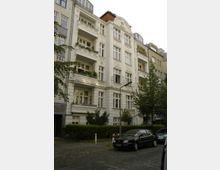 Fünfgeschossiges Wohngebäude im historischen Stil mit weißer Fassade, dekorativen Balkonen und Verzierungen. Vor dem Gebäude befinden sich geparkte Autos, ein Gehweg und Bäume.