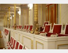 Innenraum eines historischen Opern- oder Theatersaals mit weißen Sitzreihen, roten Polstern und goldenen Verzierungen. Der Bereich ist von reich verzierten, goldenen Säulen und Wandpaneelen mit Stuck und Marmoroptik umrahmt.
