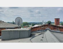 Blick über die Dächer eines Gebäudes mit Flachdach und Ziegelmauern, ausgestattet mit Satellitenschüsseln und Antennen. Im Hintergrund sind Bäume, eine Brücke und eine weite Landschaft unter einem bewölkten Himmel zu sehen.