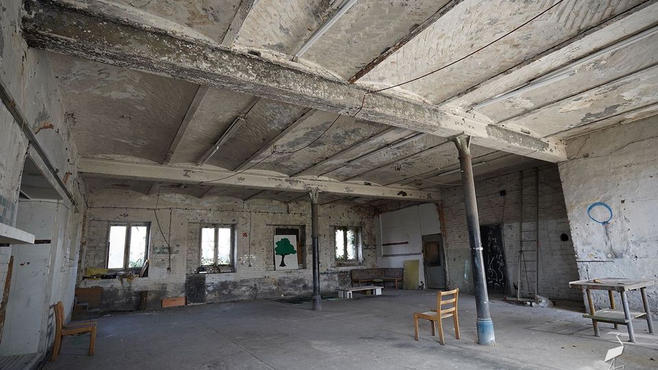 Großer, leerer Innenraum einer verlassenen Industriehalle mit roher Betondecke, freiliegenden Trägern und Stützsäulen sowie abblätternder Farbe an den Wänden. Mehrere Fenster lassen Tageslicht hinein; vereinzelt stehen einfache Stühle, ein Tisch und eine Leiter im Raum.