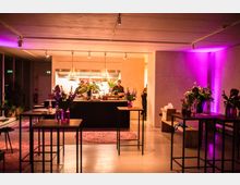 Innenraum eines modernen Veranstaltungsraums mit einer offenen Küche im Hintergrund, in der Köche arbeiten. Der Raum ist mit hohen Tischen, Blumen in Vasen und warmer, lila Beleuchtung dekoriert.