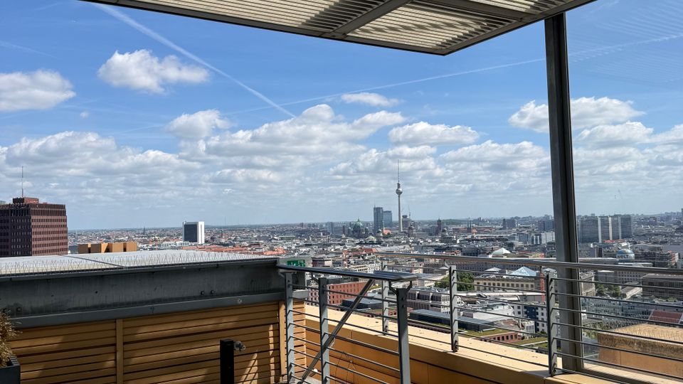 Rooftop terrace with large gray floor tiles, metal railings, and a slatted pergola roof casting striped shadows. Beyond the edge is a wide city skyline under a blue sky with scattered clouds, including a tall TV tower in the distance.
