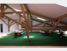 Großzügiger Dachraum mit sichtbaren Holzbalken, grünem Teppichboden und moderner Einrichtung, darunter ein Konferenztisch mit Stühlen. Tageslicht tritt durch mehrere Dachfenster ein, die weiße Wände und hohe Decken akzentuieren.