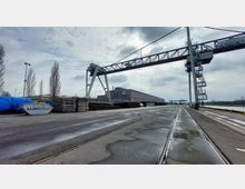 Industriehafen mit breiter Betonfläche, auf der Gleise verlaufen. Links sind gestapelte Holzplatten und ein Container zu sehen, rechts befindet sich ein großer Hafenkran. Im Hintergrund steht ein langes Lagergebäude, daneben der Wasserweg des Hafens.