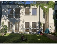 Zweistöckiges Wohngebäude mit heller Fassade und großen Fenstern, umgeben von einem kleinen Gartenbereich mit Rasen, Spielzeug, Liege und einem Holzstuhl. Ein schmaler Weg führt an der rechten Seite entlang, während Kletterpflanzen Teile der Wand bedecken.