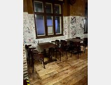 Innenraum eines einfachen Cafés oder Lokals mit Holzdielenboden, mehreren dunklen Holztischen und Stühlen sowie einem großen Holzfenster über einem Heizkörper. Die Wände sind bis zur halben Höhe weiß gestrichen und dicht mit Graffiti, Zeichnungen und Schriftzügen bedeckt.