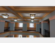 Innenansicht eines mehrstöckigen Gebäudes mit offenen Galerien, blauen Wänden und gelb-orangefarbenen Decken mit Kassettenmuster. Die Räume sind von schmiedeeisernen Geländern und großen Fenstern mit dekorativem Gitterwerk geprägt, zusätzlich hängen rechteckige Deckenlampen.