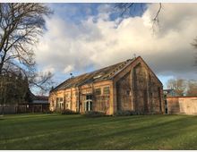 Backsteingebäude mit Satteldach in ländlicher Umgebung, umgeben von einer großen Rasenfläche und vereinzelten Bäumen. Die Fassade ist mit Kletterpflanzen bewachsen, und rechts steht ein hölzerner Schuppen.