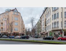 Straßenszene mit Wohn- und Geschäftshäusern, darunter eine Apotheke und ein Bistro. Im Hintergrund ist der Turm einer Kirche zu sehen, während Autos am Straßenrand parken und vereinzelte Bäume entlang des Bürgersteigs stehen.