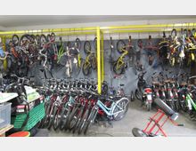 Raum mit mehreren Fahrrädern, die teils an gelben Metallgestellen an der Wand hängen und teils in geordneten Reihen auf dem Boden stehen; im Hintergrund befinden sich auch ein Motorroller und weitere Kleinteile.