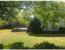 Grüne Gartenlandschaft mit einer erhöhten Holzterrasse, auf der Blumentöpfe und Terrassenmöbel stehen. Im Hintergrund ist ein beige gestrichenes Haus mit großen Fenstern und Rollläden sichtbar, umgeben von Bäumen und Sträuchern.