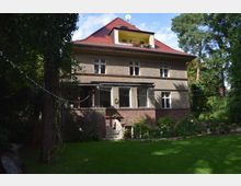 Zweistöckiges Wohnhaus mit rotem Walmdach und einer gelben Dachgaube, umgeben von einem grünen Garten mit Bäumen, Blumenbeeten und einer Rasenfläche. Eine Veranda mit Ziegelmauern und einer Treppe führt ins Haus.