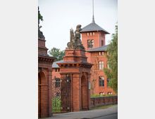 Backsteingebäude im Historismus-Stil mit einem verzierten Tor aus rotbraunem Ziegelstein und schmiedeeisernem Gitter. Im Hintergrund ist ein turmartiger Bau mit spitzem Dach und mehreren rechteckigen Fenstern zu sehen, umgeben von grüner Vegetation.
