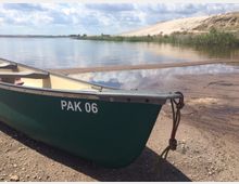 Grünes Kanu mit der Aufschrift „PAK 06“ am Ufer eines ruhigen Sees, umgeben von Sandflächen und Schilf; im Hintergrund eine flache Dünenlandschaft und bewölkter Himmel.